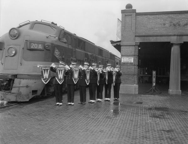 Seven U.W. Band members play " California Here We Come" on imported English fanfare trumpets decorated with a red satin Wisconsin banner. They are standing next to a locomotive at the train depot prior to a trip to Pasadena, California for the Rose Bowl game.