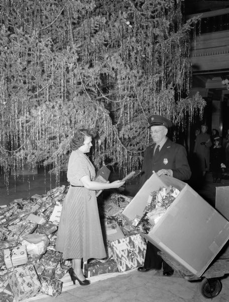 Over 2,000 children at two state institutions will receive gifts donated by state employees which are displayed under the Christmas tree in the State Capitol. The originator of the idea is Mrs. Sally Becker (left), an employee of the Bureau of Purchases; Capitol guard Millard Woodburn, is standing at right.