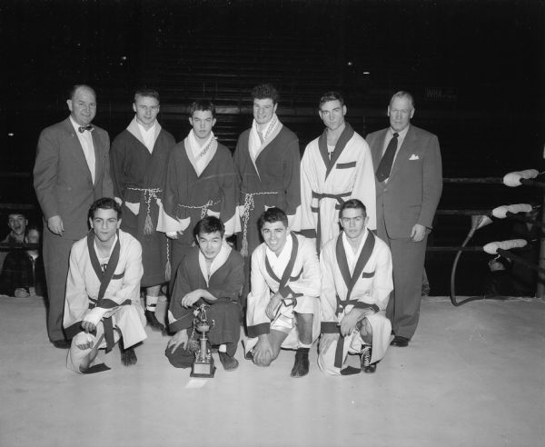 Group portrait of the winners of the UW Tournament of Contenders and their coaches, shown in the boxing ring with a trophy.  Standing, left to right: coach John Walsh, Charles Scheid, Bobby Lees, Salvatore Carling, Everett Chambers and assistant coach Vern Woodward.  Kneeling: James Greco, Roy Kuboyama, Lahaina, Bobby Goodsitt, and Lavern Lewison.