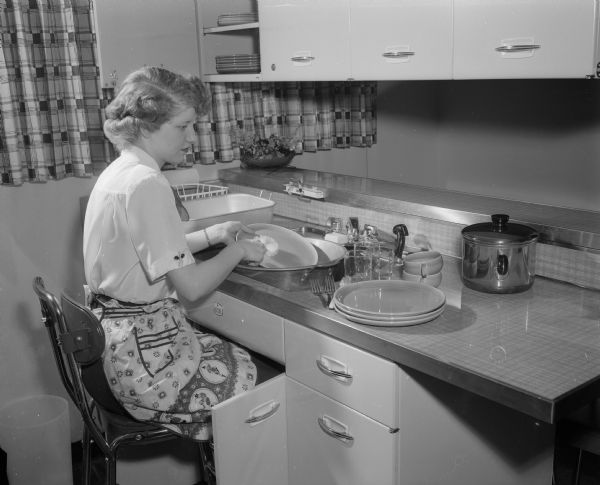 Mrs. Harry Segerstrom, Cuba City, sitting while washing dishes at a sink, demonstrating efficient dish washing methods for people with limited physical abilities at the Heart Kitchen at the Madison Vocational and Adult School.