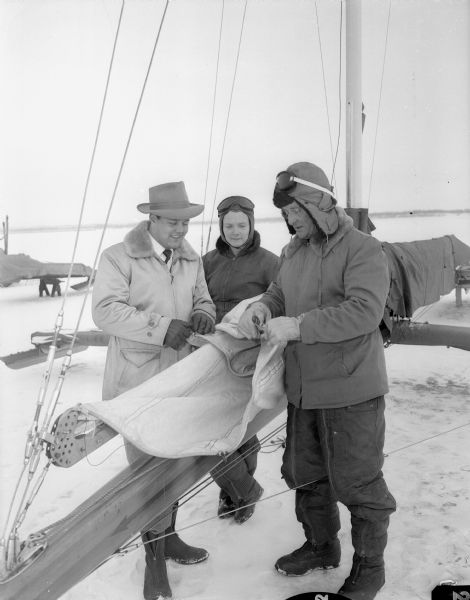 Examining the sails of the iceboat the "Mary B" are Jim Lunder, at left, and Mary Ann Bernard, center, and Carl Bernard, right. Mr. Lunder is skipper and part owner of the "Fritz," another racing boat, and Mr. Bernard sails the "Mary B." Both boats will represent Madison's Four Lakes Ice Yacht club in competition with the Oshkosh sailing club when the Hearst races being held on Lake Monona. The Madison club is the defending champion.