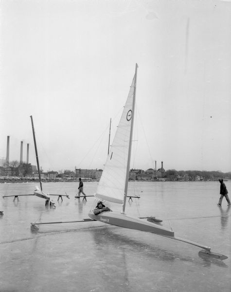 Renegade III Class E iceboat with skipper and iceboat designer Elmer Millenbach in the cockpit.  Other iceboats and the north shore of Lake Monona, including the Fauerbach Brewery, are in the background. The event was likely the Northwestern Ice Yachting Association Ice Regatta.