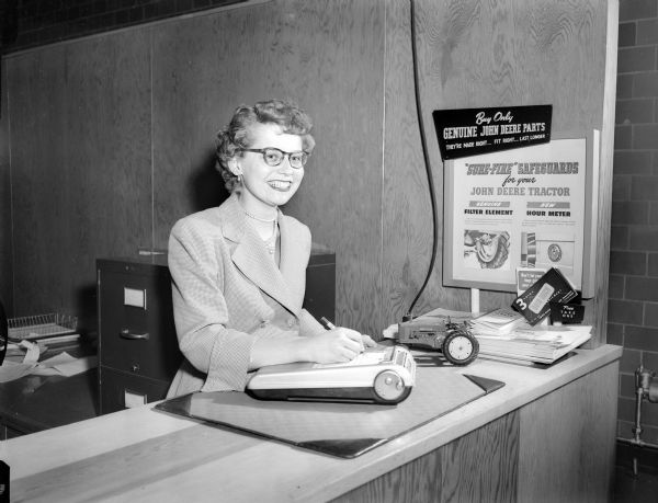 A woman stands behind a counter while writing on tablet near a sign reading "Buy Only Genuine John Deere Parts."