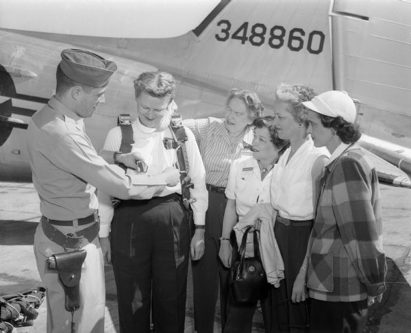 At Truax Field, Lieutenant H.B. Smith adjusts a parachute on Lillian Nitcher, one of a group of eighteen area women who were selected to make an official Air Force tour of the WAF activities at Lackland Air Force base in Texas. Waiting to be fitted with a parachute before taking off are (from left): Luella Mortenson; Mrs. Robert Marty, Monroe; Mrs. Ralph Fossahage, Mr. Horeb; and Claudia Stephan.