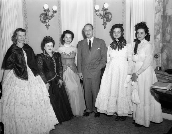 Five Prairie du Chien women dressed in period dresses and hats posing for a group portrait with Governor Walter Kohler at the Wisconsin State Capitol building. The "Sisters of the Swish" are in Madison to publicize the opening celebration of Villa Louis. Left to right: Mrs. H.J. Quartemont, Mrs. K.J. Harvey, Mrs. R.J. McWilliams, Governor Kohler, Mrs. Niles, D. Day and Mrs. Joseph Cosgriff.