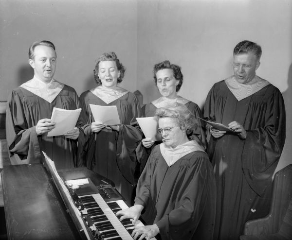 A quartet of singers wearing choir robes and holding sheet music stand and rehearse behind a seated organist, Marion Winans at First Baptist Church. From left, they are William Keith Macy, tenor; Vera Weikel Adams, soprano; Ruth Anne Piper Dykman, alto, and Ralph V. James, baritone.