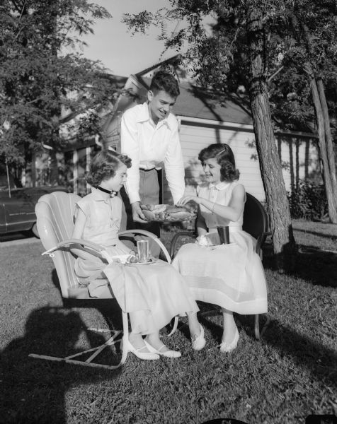 Outdoor group portrait of three Nakoma School graduates at an outdoor supper party at the home of John C. Doerfer, 4105 Chippewa Drive. David Ahlgren is serving hot dogs to Annette Thatcher, left, and Patricia Chamberlain.