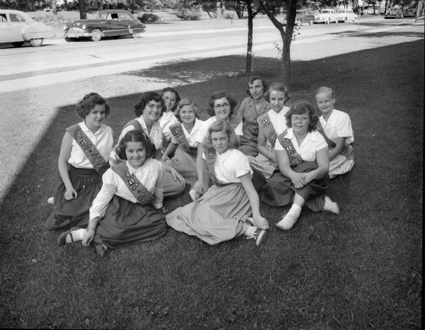 Ten seventh grade Girl Scouts of East High School Troop 48 were awarded first class badges at an outdoor ceremony on the Yahara River. They are shown wearing badge sashes over white blouses and long dark skirts. Seated from left, they are: (first row) Ruth Burzinski, Arlene Thompson, and Kirsten Flagstad, (second row) Bonnie Buchholz, Faye Fakler, Judy Ragan, Judy Oakey Helen Stone, and Lois Thomsen. In the extreme rear are Audrey Tollund and Ilene Hanson, East District girl scout director who presented the badges.