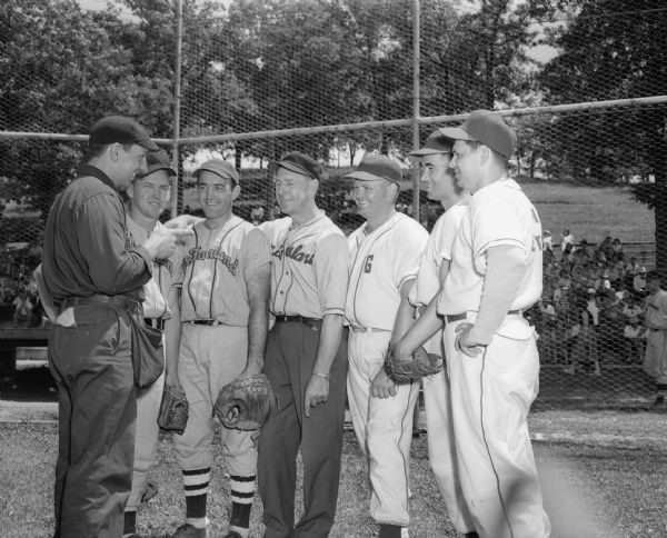 Group portrait of 6 male baseball players from different teams talking to an umpire.