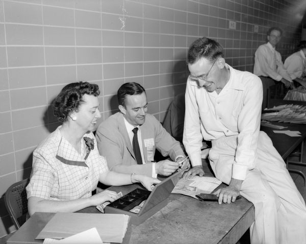 Catherine Clayton (left) is handling money and the  cash box, while Ernest Corley (center) is examining identification at the beginning of a three-day conference of the American Dairy Science Association. On the right is one of the 1,700 dairy research men, Keith Huston of the Virginia Poly-Technical Institute in Blacksburg, Virginia, dressed all in white and sitting on the table.
