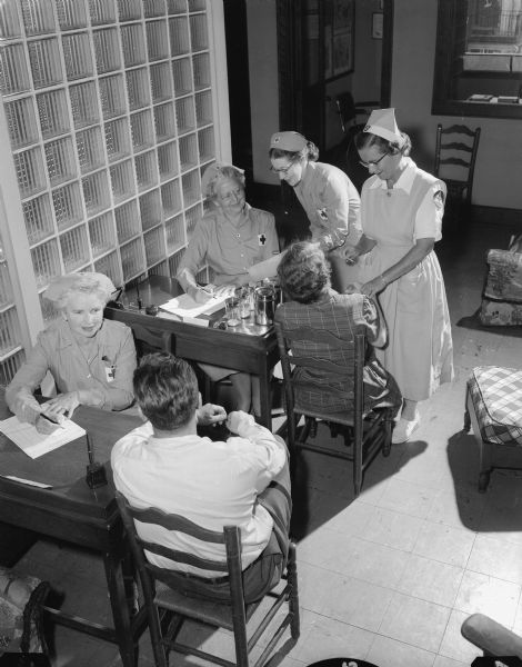 Volunteer workers of the American Red Cross, Dane County branch, assist with the routine of the blood donor program. In the foreground, Mrs. A.W. (Irene) Peterson takes a history of a blood donor. With another donor in the background, Mrs. N.J. (Madeline) Stoneman reads a thermometer. Next to her are Mrs. Myron (Gertrude) Glassen (seated) and Mrs. Adolf Soucek.