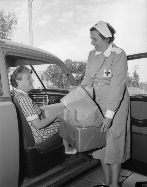 The Gray Ladies (volunteers) of the Red Cross do shopping as part of their hospital duties. Mrs. Tracy W. (Anne) Allen is shown in her car, returning from a shopping trip. Receiving the packages is Mrs. George (Deborah) Sherman.