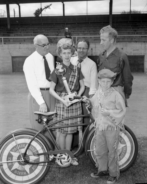 Two of the top winners in the annual youngsters' bicycle parade. Holding the trophy for the best decorated bike and best costume is Mary Coster, shown with the bicycle she won. At right is Jeffery Lynch, winner of the slogan contest. His slogan was "If you care to keep riding, keep riding with care." In the background are left to right: R.C. Salisbury, safety director of the State Motor Vehicle department; Gene Rankin, master of ceremony for the program; and Robert Reger, representing the sponsoring Montgomery Ward Company.