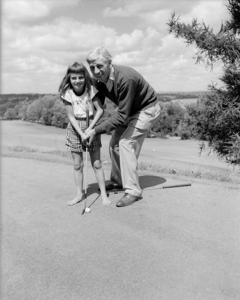 Blackhawk Country Club pro, Kully Schlicht gives a golf lesson to his daughter, Joan Schlicht, who is barefoot. In the background is a fairway, woods and a sky with clouds.