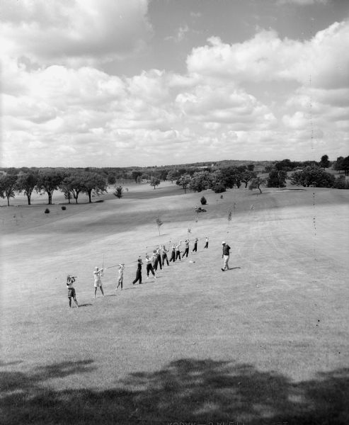 Elevated long shot view of 13 young golfers practicing their driving stance as the golf pro is looking on.  