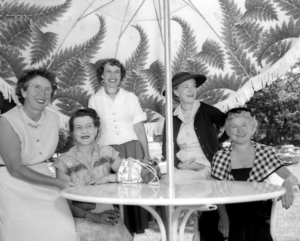 Five social chairmen from the three area country clubs sitting and standing around a table at Maple Bluff Country Club during the women's inter-club golf match. Left to right are: Edith Nystrom of Maple Bluff, Edna Walker of Maple Bluff, Reta Smith of Nakoma, Marion Pecher of Blackhawk, and Mrs. R.W. Dawley of Nakoma.
