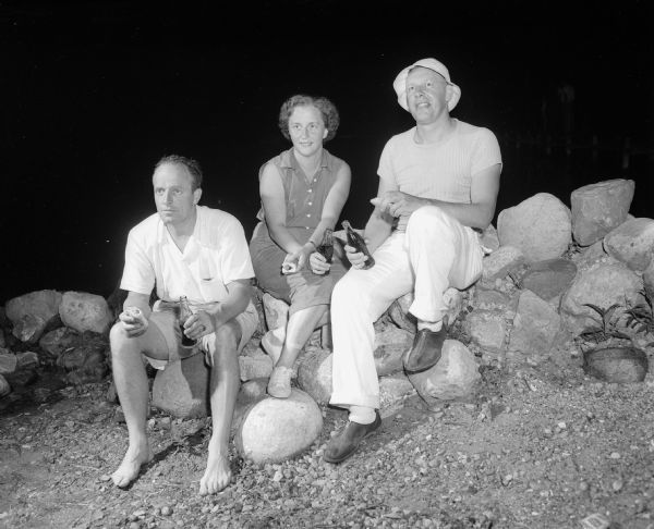 Attending the Mendota Yacht Club beachcombers party are, left to right: Philip and Dorothy Sawin, and John Esch who are enjoying a snack while sitting on rocks bordering the shoreline at the Maple Bluff beach.