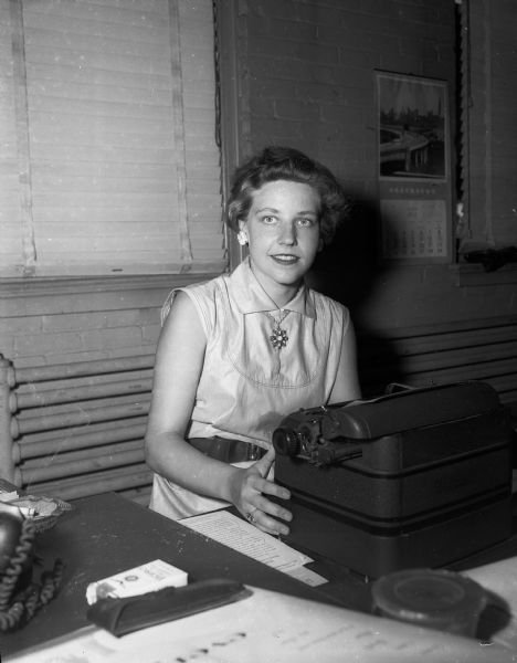 Portrait of a young woman seated at a typewriter with cigarettes, glasses case, and ash tray nearby and a Greyhound calendar on the wall. Noted to be a Dutch reporter.