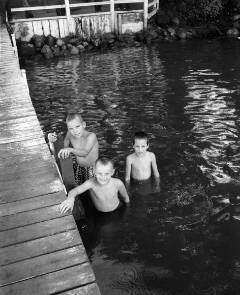 A scene from YMCA's day camp at Camp Wakanda, located on the shore of Lake Mendota.  Left to right: John Kaiser, David Williamson, and Mark Foster, standing in the water next to a pier, are taking a break from swimming.