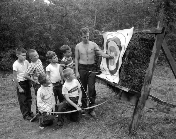 A scene from YMCA's day camp at Camp Wakanda, located on the shore of Lake Mendota.  Archery counselor, William A. Galster, checks arrows on a target with six boys gathered around him.  Foreground, left to right: Eric Whitesell and Bob Parker.  In back: David Forsyth, Mike Grans, Pat Eberlein and Philip Stansly.