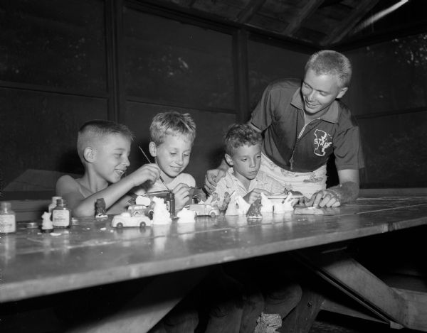 A scene from YWCA's day camp at Camp Wakonda, located on a shore of Lake Mendota. During craft time, Gerald Peck, James Foster, and Russell Bright paint small model cars and animals. Looking on is craft instructor and U.W. student Richard Thiel.