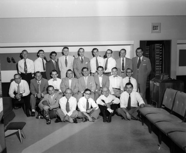 Twenty-two shoe salesmen pose for a group portrait in a store shoe department.
