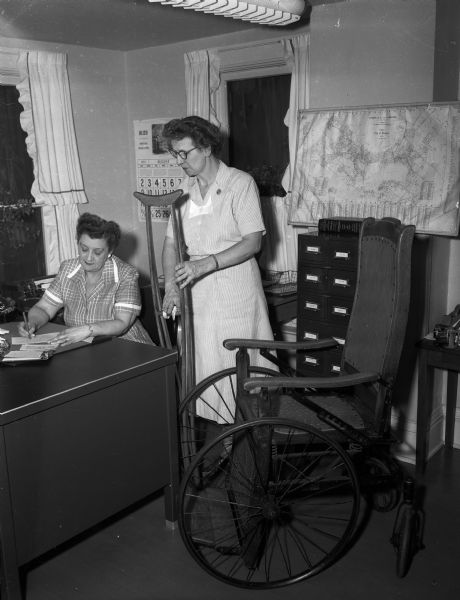 Mrs. Marjorie Southwick, secretary of the Visiting Nurse Service, checks out a pair of crutches and a wheelchair to Miss Agnes Griffith, assistant director of the agency. The agency established this loan closet to provide medical equipment needed in home care for the sick.