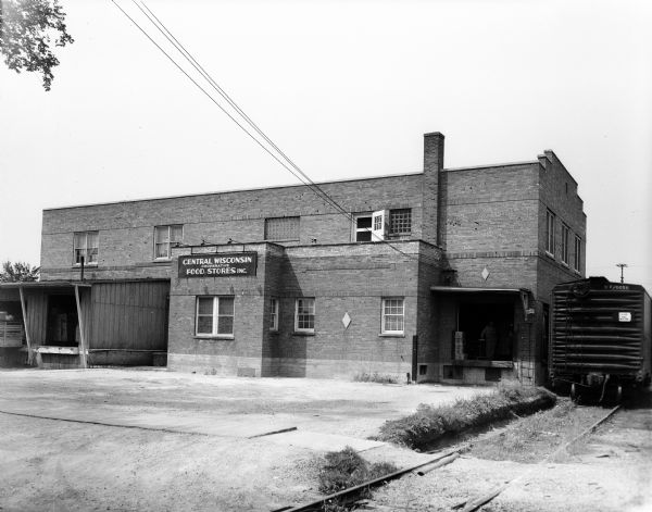 Exterior view of the Central Wisconsin Cooperative Stores, Inc. building at 112 North Lake Street. The two-story brick warehouse has four loading dock doors, and there is a railroad boxcar on the right side of the building. A man is standing in one of the doorways.