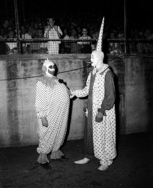 Two clowns wearing polka-dotted costumes are cavorting in front of crowded bleachers at Breese Stevens Field. The staged slow-pitch softball game was in its tenth year and drew almost 11,000 spectators on a Wednesday night. The clown at left is Steve Slattery and beside him is Leo Lacy.