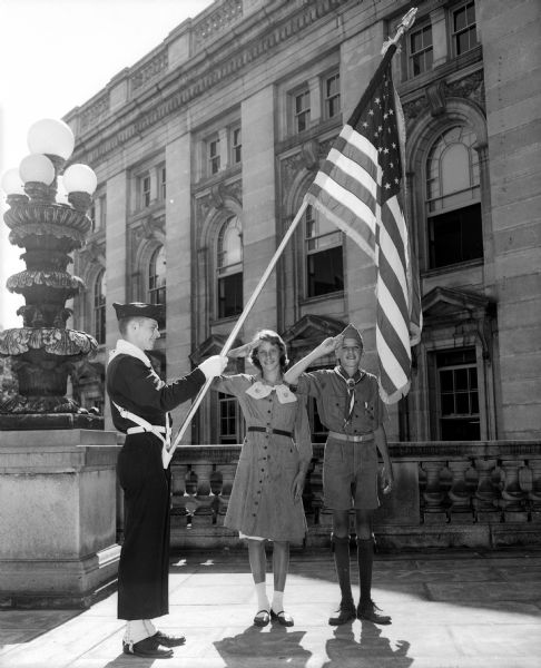 Portrait of three scouts who will participate in the Americanization Pageant. David Ross (left), member of the Boy Scout Drum and Bugle Corps, holds the American flag as Girl Scout Mary Brandt and Boy Scout William Kaeser salute. They stand on the balcony of the Monona entrance to the State Capital.