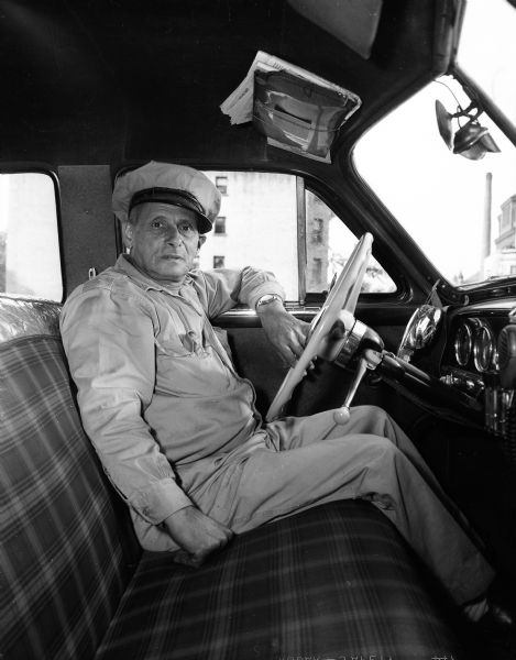 Alfred Hopf, a German Jewish refugee, wearing his uniform and sitting in his taxi, says driving a taxi in Madison is a far cry from his old career asa  prominent lawyer and banker in Germany.
