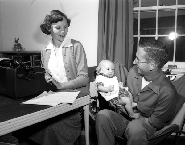 Beth Ross is sitting at a typewriter while her husband, John E., is holding their two-month-old son, Tommy. Beth is the president of the Junior Division of the University League. The Junior Division is a support organization for U.W. faculty women and wives of faculty members.