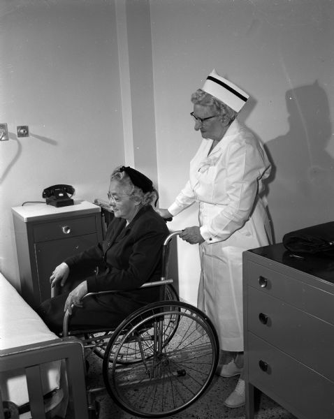 Nellie A. Brown, night supervisor of nurses, represents a modern nurse as she is pushing a patient in a wheelchair. Posing as the patient is Geneva F. McKay. The enactment is an activity during National Business Women's Week which is being observed with special features in the press, radio, and television stations to show the progress which women have made through the years. The project is sponsored by the Madison Business Women's and Professional Women's organizations.