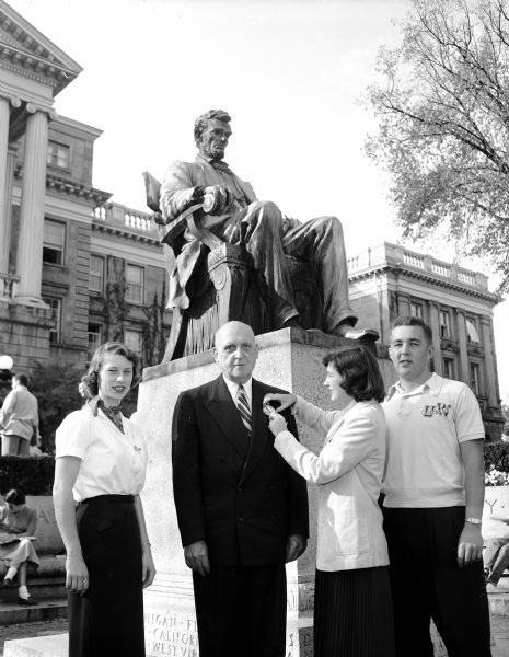University of Wisconsin President E.B. Fred is presented with the first Homecoming button by University yearbook-designated 1953 Badger Beauty, Marge Hesse. Tashia Frankfurth, co-chairman of button sales, is standing at left. At right is Fred Krull, co-chairman of button sales. Bascom Hall and the Lincoln statue are in the background.