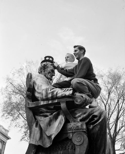 Two students perch on the lap of the seated Abraham Lincoln statue as they place a University of Wisconsin Homecoming button-decorated top hat on the figure.