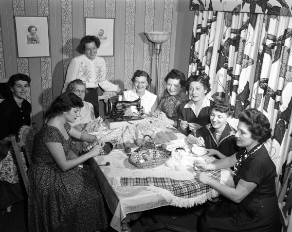 Nine women are sitting around a table while sewing in preparation for the Beth Israel sisterhood's annual bazaar at 1406 Mound Street.