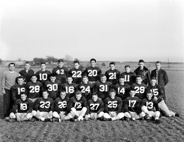 Group portrait of the Waunakee High School football team after winning the 1953 Tri-County League championship with a perfect record. Coach Dick Trotta poses with the team members, managers and assistant coach.