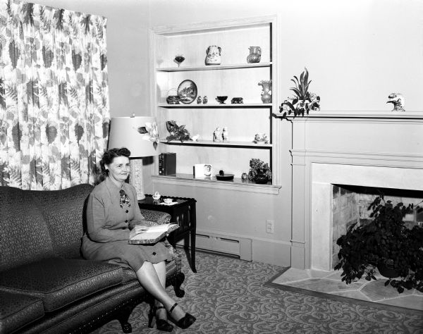 Mrs. Scheberle sitting in the living room of her newly-built home where she resides with her husband, Leo. The room features a built-in shelf, a fireplace, sofa, and other furnishings.