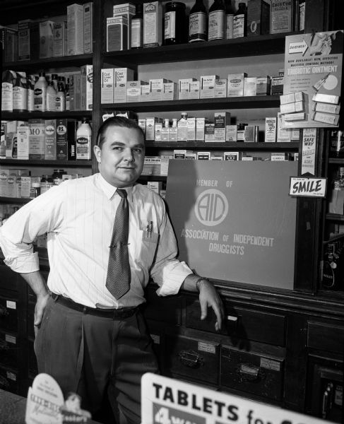 Portrait of Dick Piechowski standing in Branton's Pharmacy, 2042 East Washington Avenue, in front of a sign that reads: "Member of AID Association of Independent Druggists."