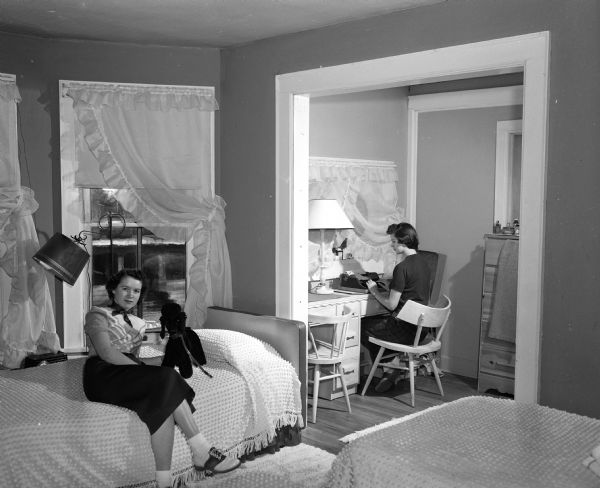 Donna Lee Genunzio of Racine works at a typewriter as Dixie Joiner of Gilman relaxes on a bed in a suite at the Ellsworth House, a residence for UW co-eds. The house is located at 422 North Murray Street.