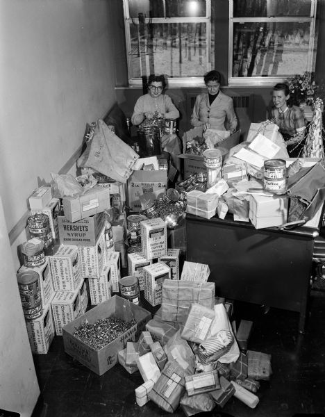 Three unidentified women (employees or volunteers) possibly at and possibly from radio station WIBA, making up Christmas gift packages of candy, nuts, and toys, to be given to homeless children. Boxes and cans with the Butternut Coffee label are piled in the foreground; these were probably donation containers used to collect the Butternut Coffee labels which were part of the collection campaign promoted by station WIBA.