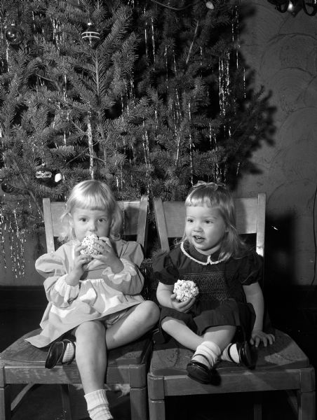 Abby Brogden, age 3, and Ashley Moffat, age 2, hold popcorn balls while they sit on chairs in front of a Christmas tree.