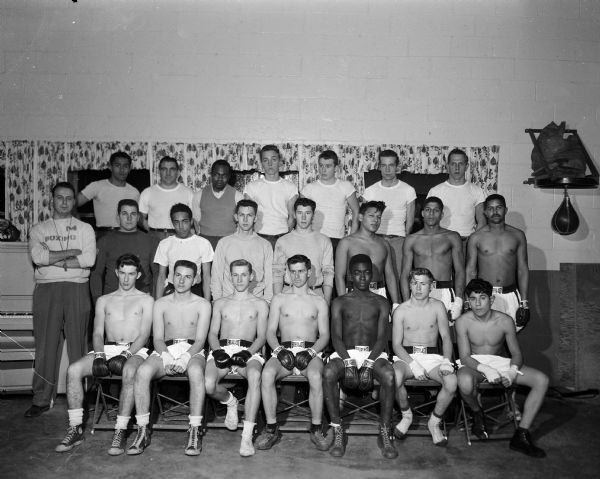Indoor group portrait of 18 of the 20-member boxing squad from St. Martin House of Madison prior to their competition at the Kenosha Golden Gloves tournament in February 1954. Also shown is the coaching staff, including Athletic Director George Holmes, and trainers Steve Badona, Lounza Adair, Adel Johnson, and Syl Farris.
