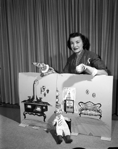Merrillyn (Mrs. Robert) Wegner is shown standing behind a set from "Your Puppet Pals" which includes 3 puppets: Professor Bookworm, Dingle the Clown, and Fuzzywig. "Your Puppet Pals" was a weekly show on WKOW-TV. Merrillyn was known in later life as Lynn Hartridge.