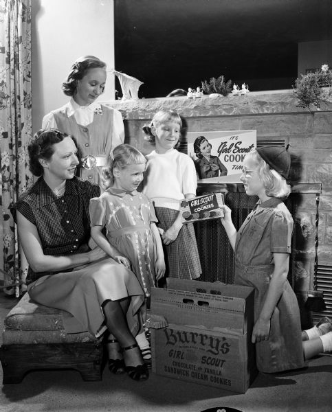 Mrs. William C. Roberts (left) of 29 Frederick Circle, co-chairman of the annual Girl Scout cookie sale in Madison, posing with her four daughters, Blanche, 13, Bessie Jean, 4, Kathleen, 7, and Beverly Ann, 10 (wearing her Brownie uniform). The group portrait includes their fireplace, a large Burry's cookie box and a small girl scout cookie box. There is also a poster on the mantle stating: "It's Girl Scout Cookie Time."
