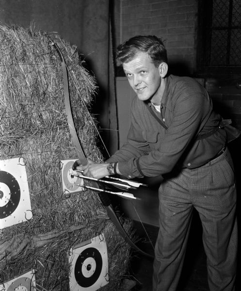 A man is standing with bow and arrows at the target of the first annual invitational indoor archery meet held at the YMCA range. He is probably George Van Tassel, president of the host Blackhawk Bow Hunters Club, winner of the men's instinctive shooting event.