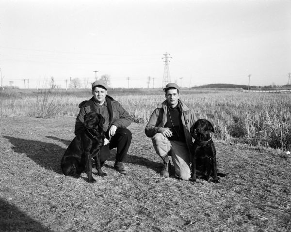 Two trainers posing outdoors with their dogs after taking part in the Madison field trials. A record number of 80 dogs were entered. Winners in the four categories (all black labradors) were "Smokey," owned by Irv LaSage of Wauwatosa in the open all-age class; "Cinders of Erin," owned by J. Detienne of Milwaukee in the derby class; "Mickie," owned by Jim McAndrews of Decorah, Iowa in the amateur trained and handled class; and "Dela Winn's Desraeli," owned by Irv Duchner of Milwaukee in the qualifying class.