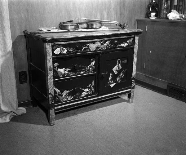 Music cabinet with decoupage design by Fern Fowler. The cabinet was owned by Winfield & Irene Alexander; a violin is laying on top of the cabinet.