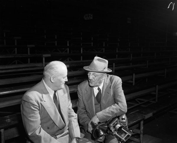 Harry Conley, left, Director of Athletics in the Superior school system, and Arthur M. Vinje, Wisconsin State Journal photographer, sitting on the bleachers at the state high school basketball tournament while reminiscing over old times. They were boyhood chums in Superior. Vinje is holding his 4 x 5 press camera.