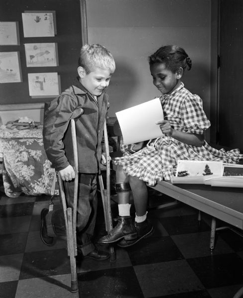 Terry Snowden stands with crutches and looks on as Marjorie Parish, seated and wearing a leg brace with a crutch at her side, examines a pamphlet. They are first grade pupils at Washington Orthopedic School at 545 West Dayton Street. They hope to attend Camp Wawbeek, a summer camp sponsored by Easter Seals.